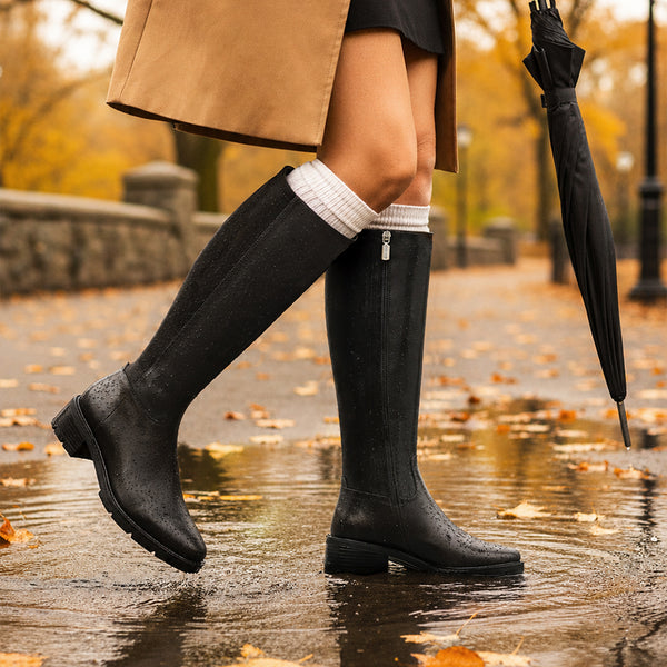 HARRIET A person walking in a wet park path wearing tall black rain boots, with a tan coat, white socks, and a black umbrella visible against an autumn background. - thumbnail image 2 of 21