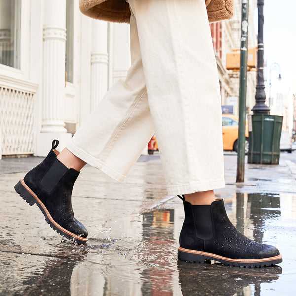 MAYES A person wearing waterproof black suede Chelsea-style ankle boots with elastic side panels and tan soles, walking on a wet city street with puddles. The person is also wearing off-white wide-leg pants. - thumbnail image 2 of 54