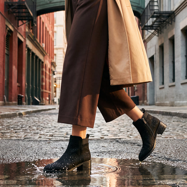 SHYNE A person walking on a cobblestone city street wearing black ankle boots with silver rhinestone embellishments, brown wide-leg cropped pants, and a beige trench coat, stepping into a puddle. - thumbnail image 2 of 31