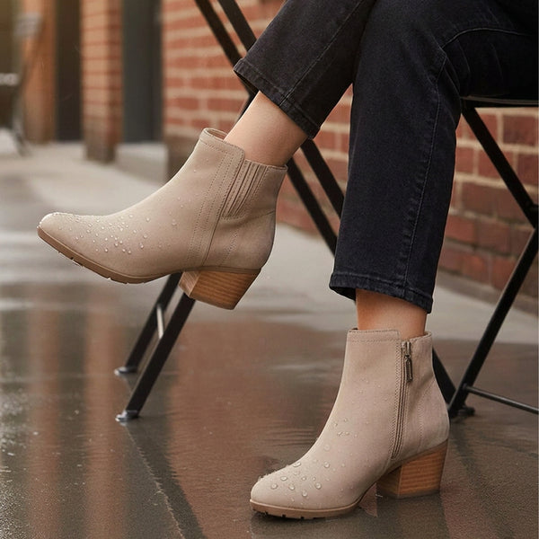 VALLI2.0 A person seated on a chair wearing waterproof beige suede ankle boots with a low block heel, side zipper, and small silver details, photographed outdoors on a wet street with brick buildings in the background. - thumbnail image 2 of 54